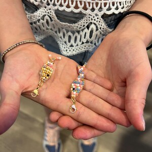 May include: Two small fish-shaped charms crafted from tiny multi-colored glass beads. One charm features yellow, white, and pink beads, while the other displays a mix of blue, pink, and white beads. Each charm includes a gold-toned metal lobster clasp.