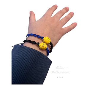 May include: Two handmade bracelets with yellow Lego-style heads and black and blue woven cords. The bracelets are on a wrist, with the blue bracelet on top. The heads have smiling faces.