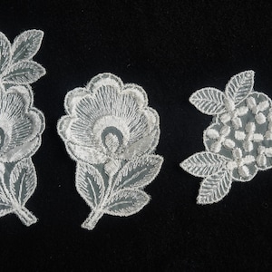 Könnte beinhalten: Drei weiße Spitzenblumenapplikationen mit zarten Blumendesigns. Die Applikationen sind perfekt, um Kleidung, Kunsthandwerk oder Wohnaccessoires einen Hauch von Eleganz zu verleihen.