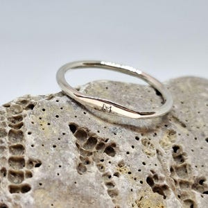 Op de afbeelding: Een zilveren ring met een minimalistisch design. De ring heeft een gladde, gepolijste afwerking en een subtiel, gegraveerd detail. De ring rust op een getextureerde, lichtgrijze steen, waardoor een contrast in texturen en tinten ontstaat. De eenvoudige elegantie van de ring maakt het een veelzijdig accessoire.