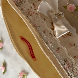 K&ouml;nnte beinhalten: Eine wei&szlig;e Rei&szlig;verschlusstasche mit Blumenmuster und Spitzenbesatz. Eine wei&szlig;e Satinschleife mit einem silbernen Anh&auml;nger ist oben angebracht. Die Tasche ist offen und das Innere ist mit einem hellgelben Stoff ausgekleidet. Der Text "Cozy Times" ist in Rot auf einem wei&szlig;en Etikett im Inneren der Tasche aufgedruckt.