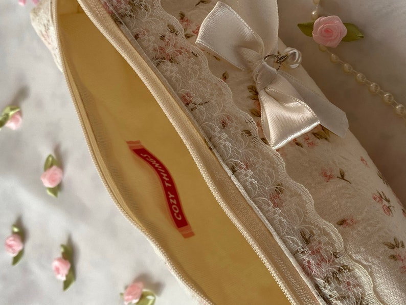 K&ouml;nnte beinhalten: Eine wei&szlig;e Rei&szlig;verschlusstasche mit Blumenmuster und Spitzenbesatz. Eine wei&szlig;e Satinschleife mit einem silbernen Anh&auml;nger ist oben angebracht. Die Tasche ist offen und das Innere ist mit einem hellgelben Stoff ausgekleidet. Der Text "Cozy Times" ist in Rot auf einem wei&szlig;en Etikett im Inneren der Tasche aufgedruckt.