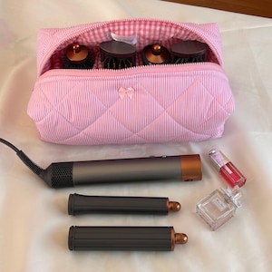 May include: A pink and white striped quilted cosmetic bag with a bow detail, containing hair styling tools. A hair dryer, two styling attachments, lip gloss, and perfume are also visible.