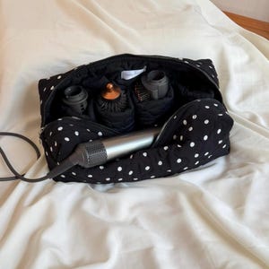 May include: A black quilted travel case with a white polka dot pattern, featuring a silver zipper closure. Inside, the bag contains a metallic gray hair dryer with a long cord and three distinct hair styling attachments organized in compartments.