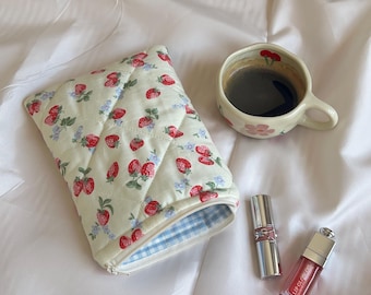 May include: A cream-colored quilted pouch with a strawberry and floral pattern, a cup of coffee, and two tubes of lip products. The pouch has a blue and white gingham lining and a zipper closure. The cup is decorated with flowers.