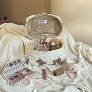 May include: An open, quilted cosmetic bag with a teddy bear pattern, filled with makeup and a small teddy bear. A palette of eyeshadows, lipstick, and a mug with a teddy bear design are also present. The bag has the text "cozy things".