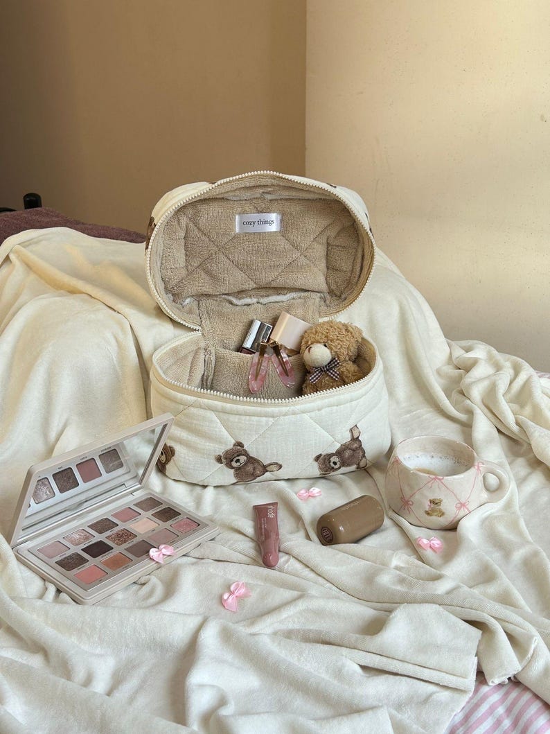 May include: An open, quilted cosmetic bag with a teddy bear pattern, filled with makeup and a small teddy bear. A palette of eyeshadows, lipstick, and a mug with a teddy bear design are also present. The bag has the text "cozy things".