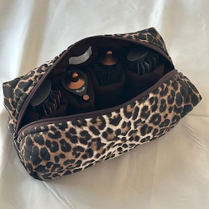 Può includere: Borsa per cosmetici aperta con stampa leopardata, cerniera e interno marrone. La borsa contiene strumenti per lo styling dei capelli, tra cui spazzole e un asciugacapelli. La borsa è trapuntata e ha il testo "cozy dune" all'interno.