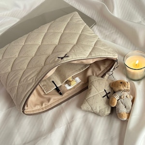 May include: Beige quilted laptop sleeve with black dot pattern, open to reveal a beige interior. A matching small pouch with a black cross design and a teddy bear keychain are next to the sleeve. A lit candle sits nearby.