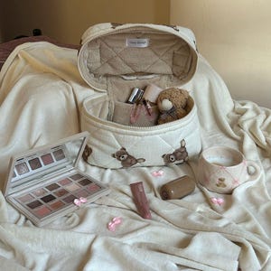 May include: An open, quilted makeup bag with teddy bear designs, containing perfume bottles and a small teddy bear. A makeup palette, lipstick, and a mug with a teddy bear design are also present. The bag has the text "cozy things".