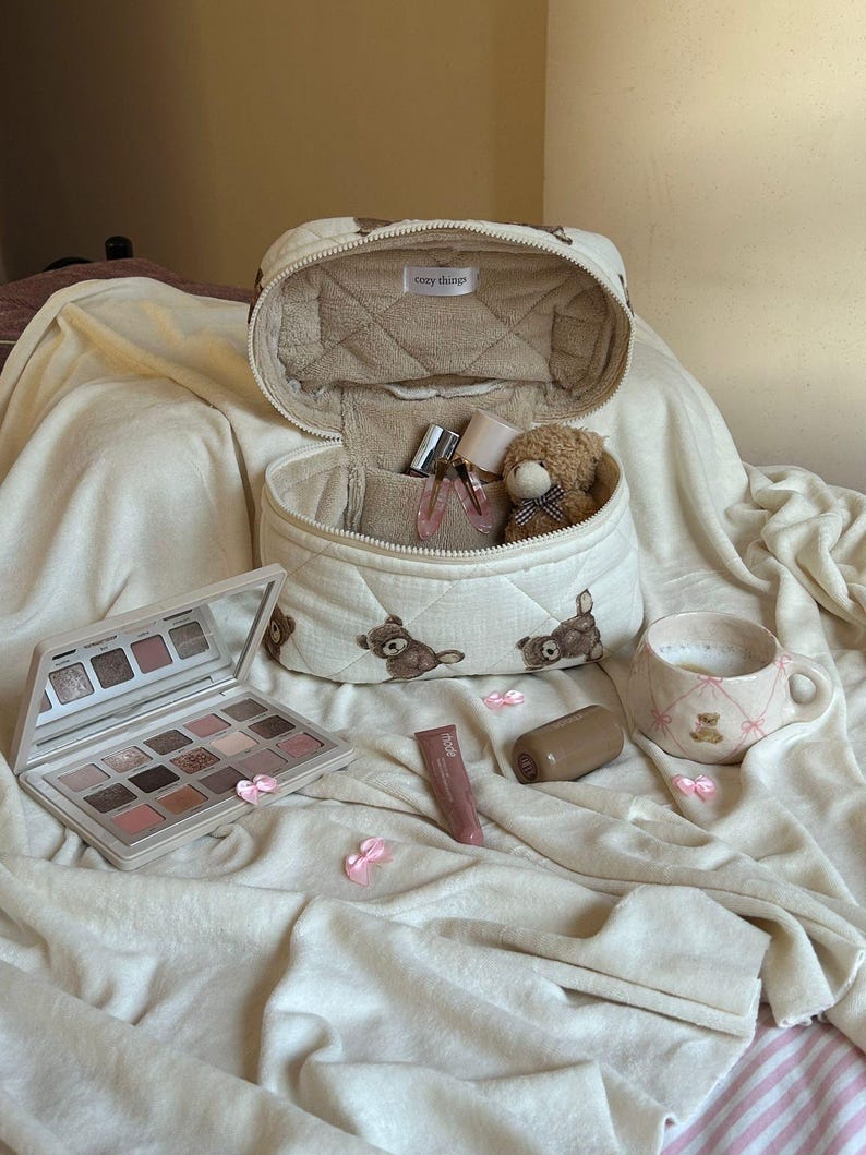 May include: An open, quilted makeup bag with teddy bear designs, containing perfume bottles and a small teddy bear. A makeup palette, lipstick, and a mug with a teddy bear design are also present. The bag has the text "cozy things".