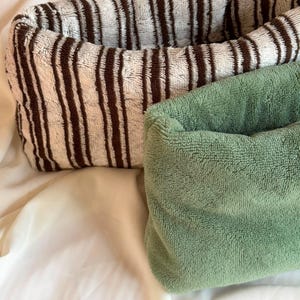 May include: Two rectangular fabric storage baskets with a soft, plush texture. One basket features a pattern of vertical dark brown stripes on a cream background, while the other basket is a solid, muted sage green color with a consistent fuzzy finish.