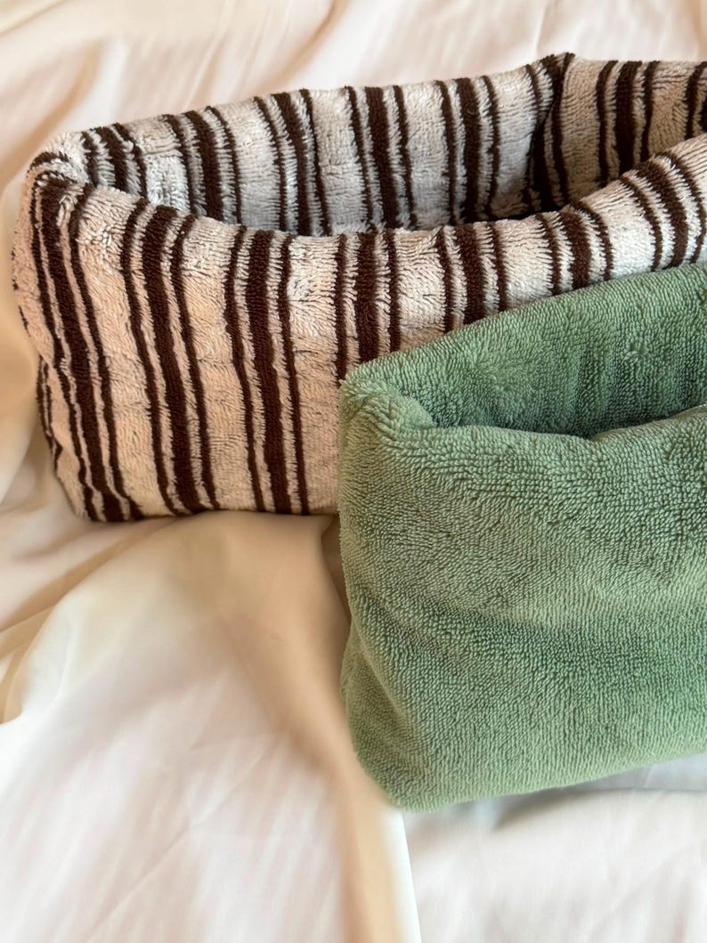May include: Two rectangular fabric storage baskets with a soft, plush texture. One basket features a pattern of vertical dark brown stripes on a cream background, while the other basket is a solid, muted sage green color with a consistent fuzzy finish.