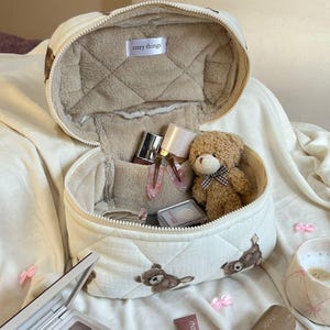 May include: Cream-colored quilted cosmetic bag with a teddy bear pattern, open to reveal makeup and a small teddy bear. The bag has a zipper closure and the text "cozy things". A makeup palette and other beauty products are also visible.
