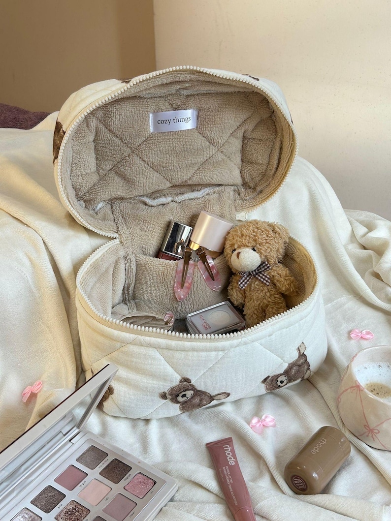 May include: Cream-colored quilted cosmetic bag with a teddy bear pattern, open to reveal makeup and a small teddy bear. The bag has a zipper closure and the text "cozy things". A makeup palette and other beauty products are also visible.