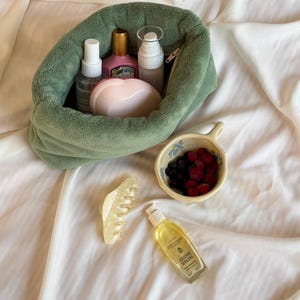 May include: A sage green plush pouch containing various skincare bottles and a pink heart-shaped compact. Beside it sits a small ceramic bowl filled with dark berries, a translucent cream-colored hair claw, and a bottle of L'Occitane Amande Sublime oil.