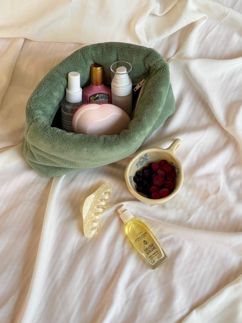 May include: A sage green plush pouch containing various skincare bottles and a pink heart-shaped compact. Beside it sits a small ceramic bowl filled with dark berries, a translucent cream-colored hair claw, and a bottle of L'Occitane Amande Sublime oil.