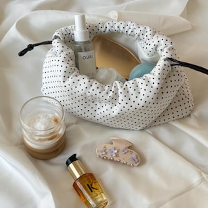 Può includere: Un sacchetto bianco con coulisse e pois neri contiene prodotti di bellezza, tra cui una spazzola per capelli e un flacone spray. Un bicchiere di caffè, una clip per capelli floreale e una bottiglia di olio per capelli sono vicini.