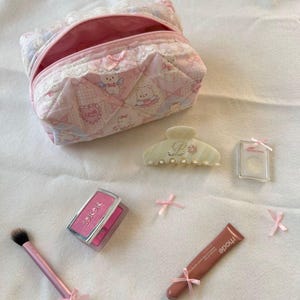 May include: A collection of beauty products, including a pink quilted cosmetic bag, blush, lip gloss, a hair clip, and a makeup brush. The items are decorated with pink bows and arranged on a white surface.