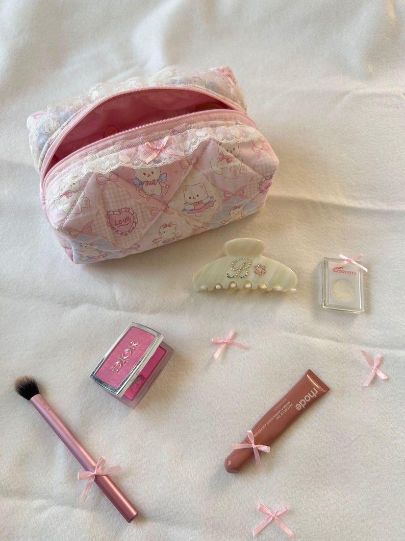 May include: A collection of beauty products, including a pink quilted cosmetic bag, blush, lip gloss, a hair clip, and a makeup brush. The items are decorated with pink bows and arranged on a white surface.