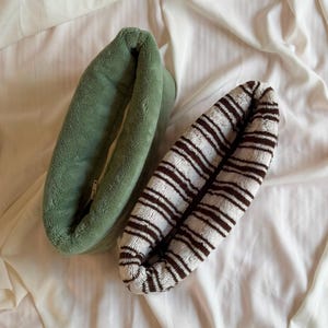 May include: Two oval-shaped, plush fabric pouches with zippered closures. One pouch is a solid sage green color, while the other features a pattern of thin, dark brown horizontal stripes on a cream-colored background, both with a soft, fuzzy texture.