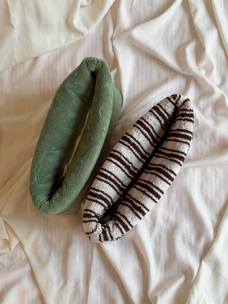 May include: Two oval-shaped, plush fabric pouches with zippered closures. One pouch is a solid sage green color, while the other features a pattern of thin, dark brown horizontal stripes on a cream-colored background, both with a soft, fuzzy texture.