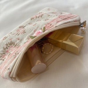 May include: A small, white zippered pouch with a floral pattern and pink embroidery. The pouch is open and contains a gold lipstick, a gold compact, and a gold chain necklace.