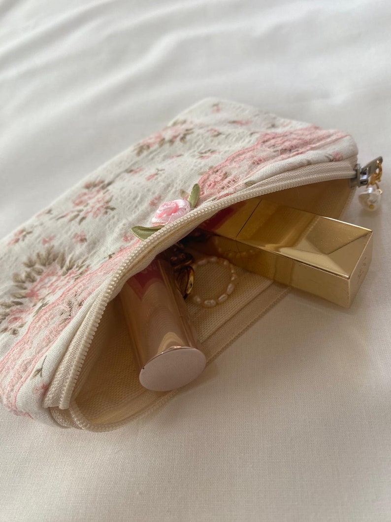 May include: A small, white zippered pouch with a floral pattern and pink embroidery. The pouch is open and contains a gold lipstick, a gold compact, and a gold chain necklace.