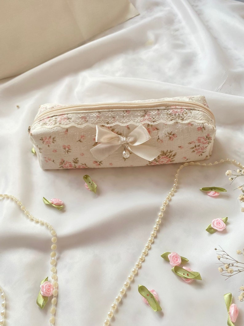 K&ouml;nnte beinhalten: Eine kleine, wei&szlig;e Rei&szlig;verschlusstasche mit Blumenmuster und wei&szlig;em Spitzenbesatz. Eine wei&szlig;e Satinschleife mit Perlenakzent ist an der Vorderseite der Tasche befestigt.