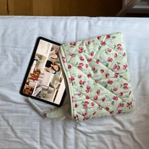 Może przedstawiać: Pikowane etui na tablet z wzorem truskawek w kolorze czerwonym i zielonym na jasnym żółtym tle. Tablet i biały przedmiot dekoracyjny leżą na białej powierzchni.