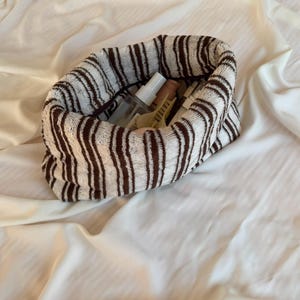 May include: A soft, cylindrical fabric pouch featuring a white and brown vertical striped pattern. The open top reveals several small personal care items, including a clear spray bottle, a beige tube, and a yellow hair clip tucked inside.