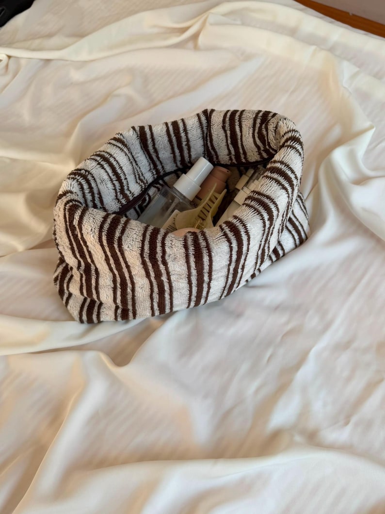 May include: A soft, cylindrical fabric pouch featuring a white and brown vertical striped pattern. The open top reveals several small personal care items, including a clear spray bottle, a beige tube, and a yellow hair clip tucked inside.