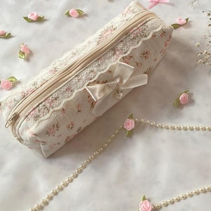 K&ouml;nnte beinhalten: Eine wei&szlig;e Stofftasche mit Blumenmuster und Spitzenbesatz. Die Tasche hat einen Rei&szlig;verschluss und eine wei&szlig;e Schleife aus Satin. Die Tasche ist von rosa Stoffblumen und einer Perlenkette umgeben.