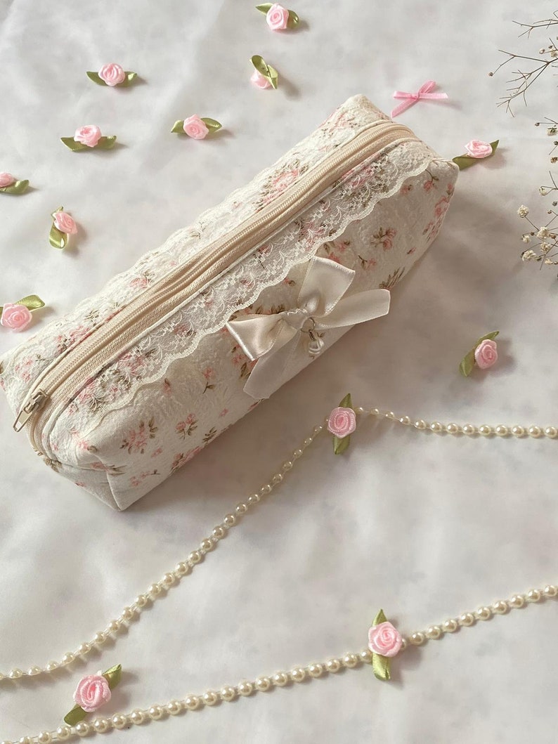 K&ouml;nnte beinhalten: Eine wei&szlig;e Stofftasche mit Blumenmuster und Spitzenbesatz. Die Tasche hat einen Rei&szlig;verschluss und eine wei&szlig;e Schleife aus Satin. Die Tasche ist von rosa Stoffblumen und einer Perlenkette umgeben.