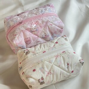 May include: Two quilted cosmetic bags. One is pink with a repeating pattern of cartoon bears, bows, and hearts. The other is white with a similar pattern. Both have a zipper closure and are trimmed with lace.