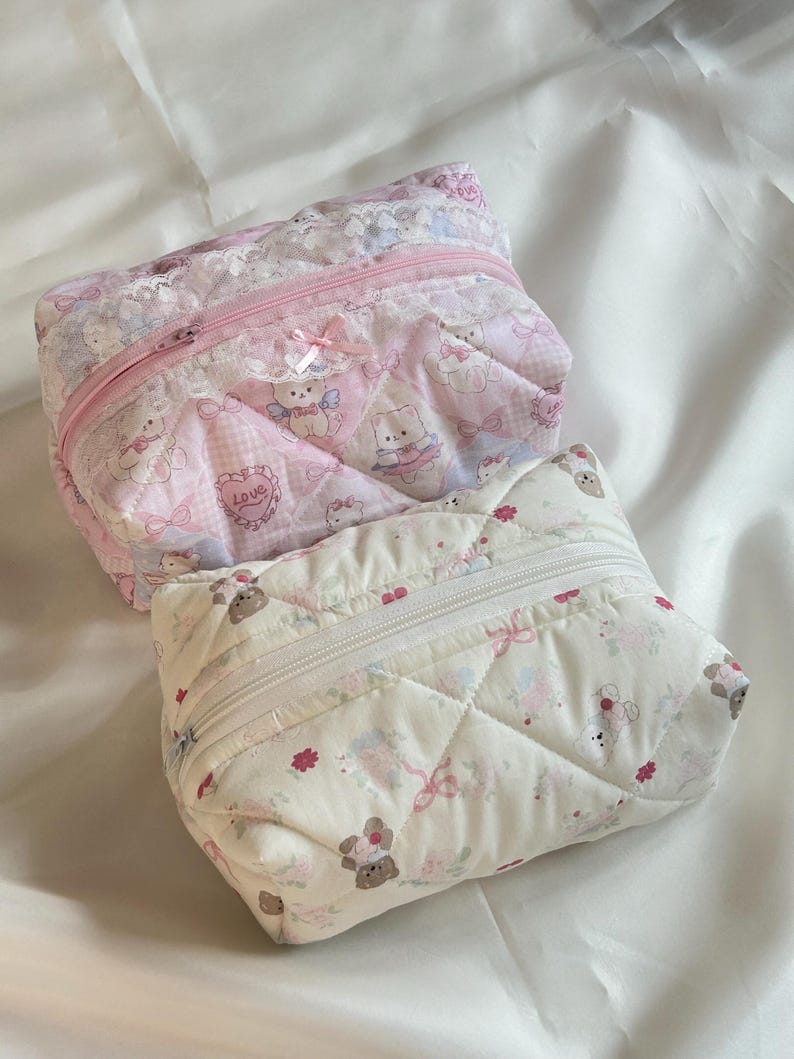 May include: Two quilted cosmetic bags. One is pink with a repeating pattern of cartoon bears, bows, and hearts. The other is white with a similar pattern. Both have a zipper closure and are trimmed with lace.