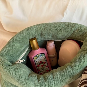 May include: A sage green terry cloth pouch containing a pink Gucci Flora perfume bottle with a gold cap, a light pink Caudalie skincare pump bottle, and a rounded light pink hairbrush. The pouch features a white zipper closure along the top.