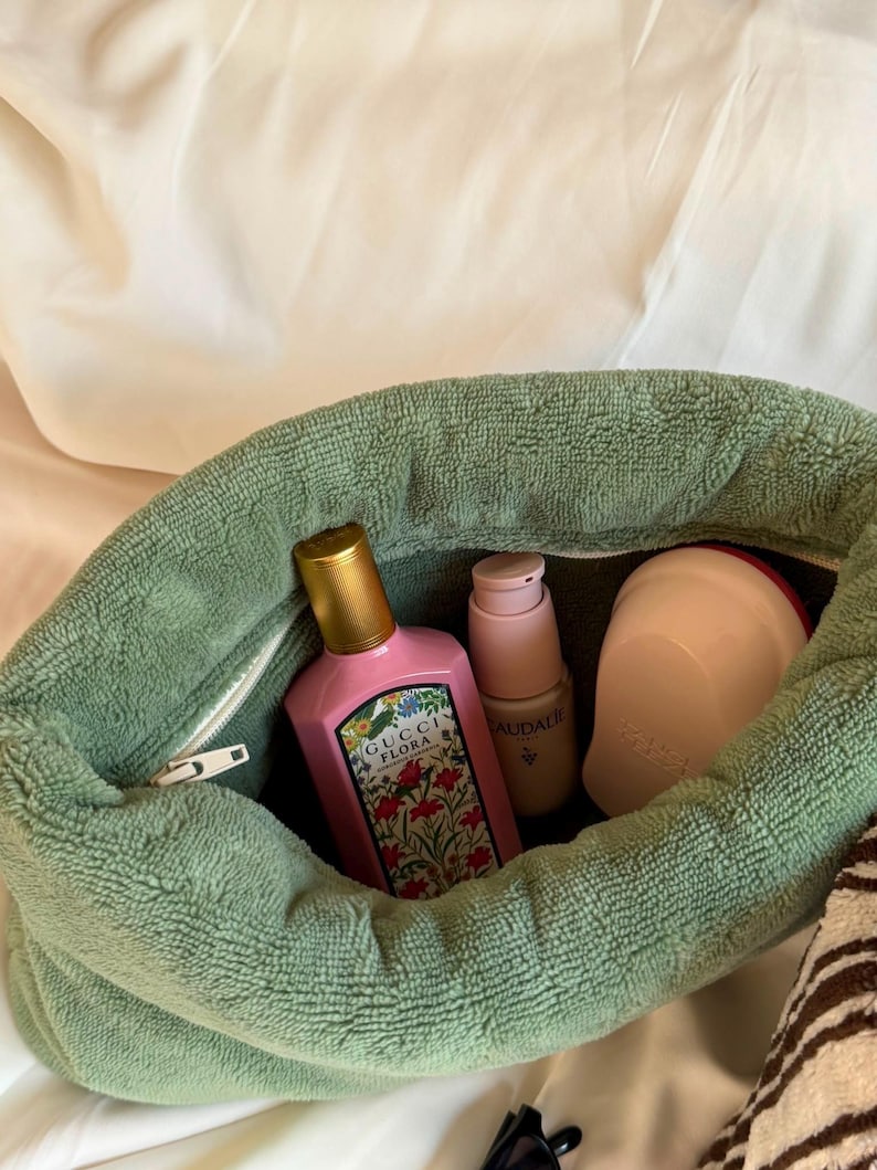 May include: A sage green terry cloth pouch containing a pink Gucci Flora perfume bottle with a gold cap, a light pink Caudalie skincare pump bottle, and a rounded light pink hairbrush. The pouch features a white zipper closure along the top.