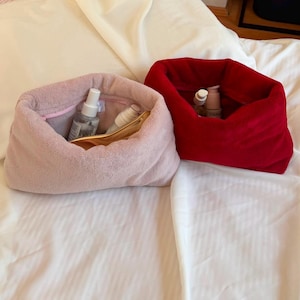 Può includere: Due morbide pochette cosmetiche in tessuto felpato, una rosa chiaro e una rosso scuro, con un'ampia apertura. La pochette rosa contiene due piccoli flaconi spray trasparenti e un pettine dorato, mentre quella rossa contiene due flaconi.