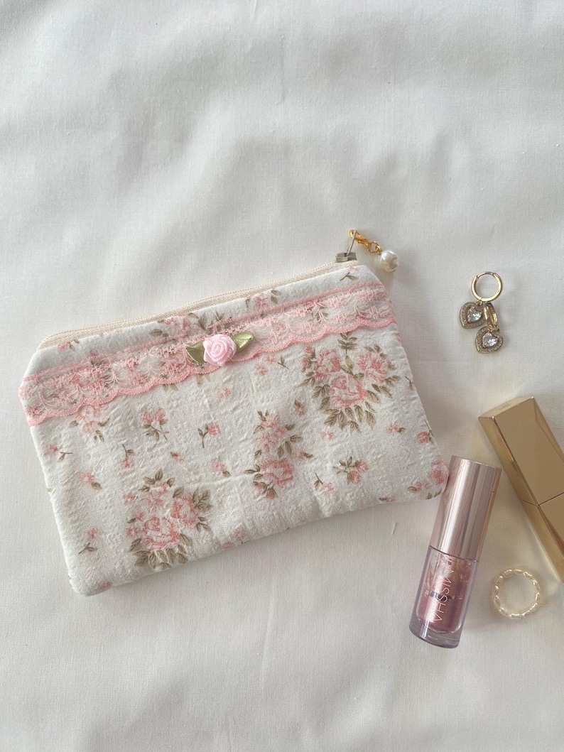 May include: A small white zippered pouch with a pink floral pattern and lace trim. The pouch has a pink rose embellishment and a gold zipper pull with a pearl charm. The pouch is sitting on a white surface with a gold and pink lip gloss tube and gold rings.