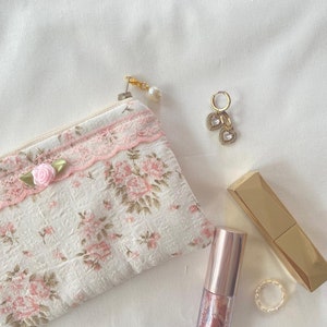 May include: A small floral print zipped pouch with a pink lace trim, a gold lipstick tube, a gold lipstick tube with a clear lid, a pair of gold heart-shaped earrings with clear stones, and a gold ring on a white background.
