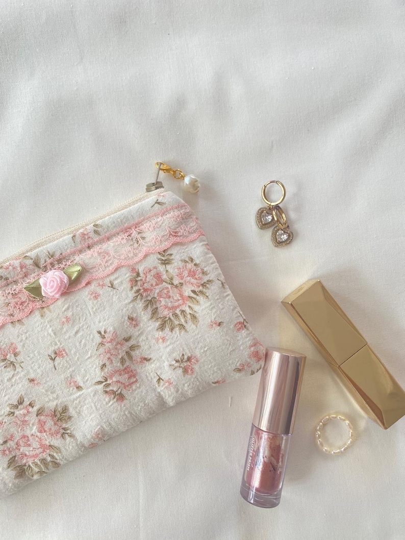 May include: A small floral print zipped pouch with a pink lace trim, a gold lipstick tube, a gold lipstick tube with a clear lid, a pair of gold heart-shaped earrings with clear stones, and a gold ring on a white background.