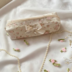 K&ouml;nnte beinhalten: Eine kleine, rechteckige, cremefarbene Rei&szlig;verschlusstasche mit Blumenmuster und Spitzenbesatz. Eine wei&szlig;e Satinschleife mit Perlenakzent ist an der Tasche befestigt. Die Tasche liegt auf einer wei&szlig;en Stoffoberfl&auml;che mit einer Perlenkette und rosa Blumenverzierungen.