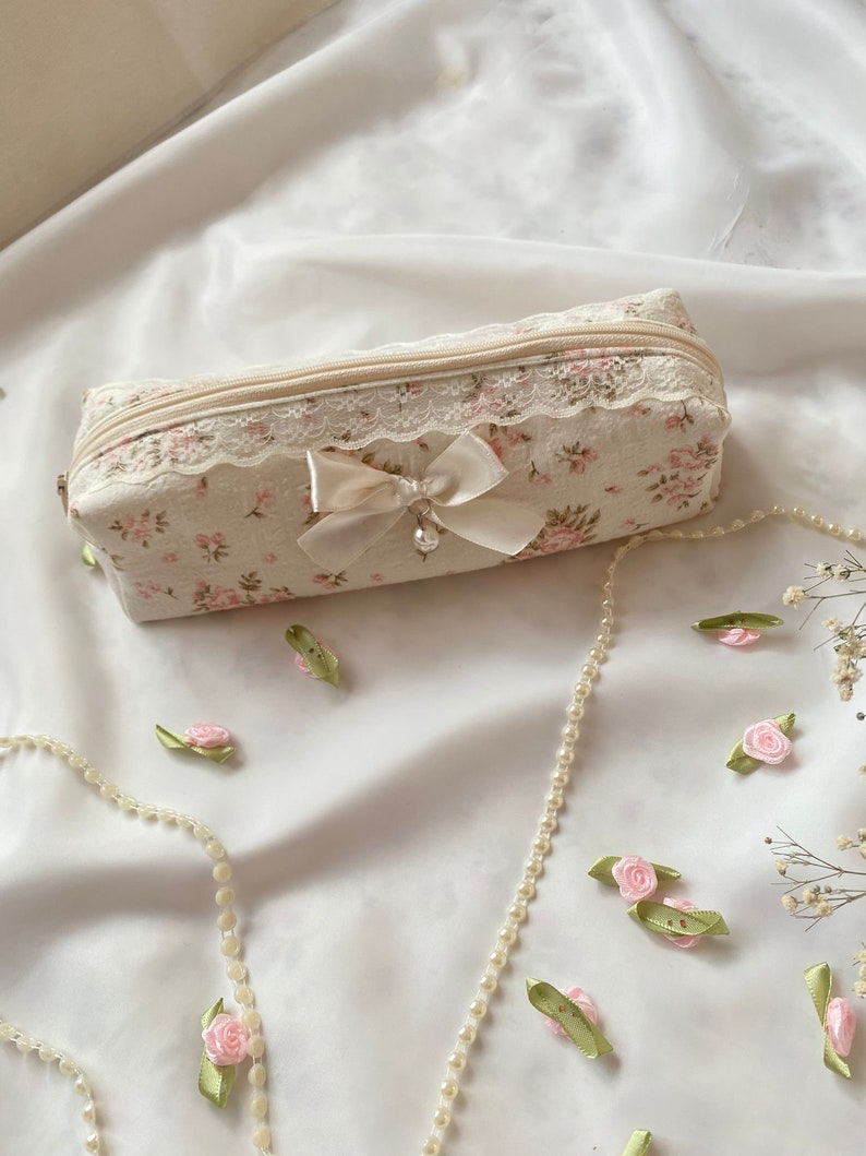 K&ouml;nnte beinhalten: Eine kleine, rechteckige, cremefarbene Rei&szlig;verschlusstasche mit Blumenmuster und Spitzenbesatz. Eine wei&szlig;e Satinschleife mit Perlenakzent ist an der Tasche befestigt. Die Tasche liegt auf einer wei&szlig;en Stoffoberfl&auml;che mit einer Perlenkette und rosa Blumenverzierungen.