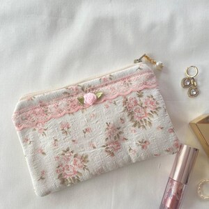 May include: A small white zippered pouch with a pink floral pattern and lace trim. A pink rose embellishment is attached to the zipper pull. The pouch is lying on a white surface.