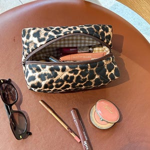 May include: A brown and black leopard print quilted makeup bag with a zipper closure. The bag is open and contains various makeup items, including a pink lipstick, a clear lip gloss, and a brown eyeliner pencil. The bag is sitting on a brown leather surface.
