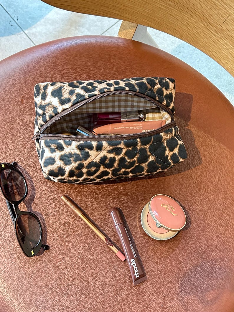 May include: A brown and black leopard print quilted makeup bag with a zipper closure. The bag is open and contains various makeup items, including a pink lipstick, a clear lip gloss, and a brown eyeliner pencil. The bag is sitting on a brown leather surface.