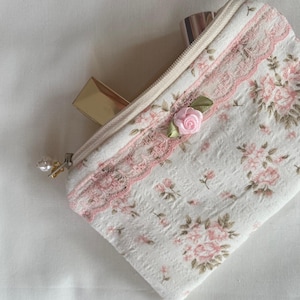 May include: A small white zippered pouch with a floral print and pink lace trim. A pink rose with green leaves is sewn onto the pouch. The pouch is open and contains two lipstick tubes.