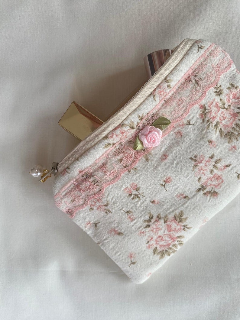 May include: A small white zippered pouch with a floral print and pink lace trim. A pink rose with green leaves is sewn onto the pouch. The pouch is open and contains two lipstick tubes.
