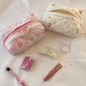 May include: Two quilted cosmetic bags, one pink and one cream, with a teddy bear pattern. Accessories include a hair clip, blush compact, eyeshadow, makeup brush, and lipstick. Pink bows are scattered around the items.